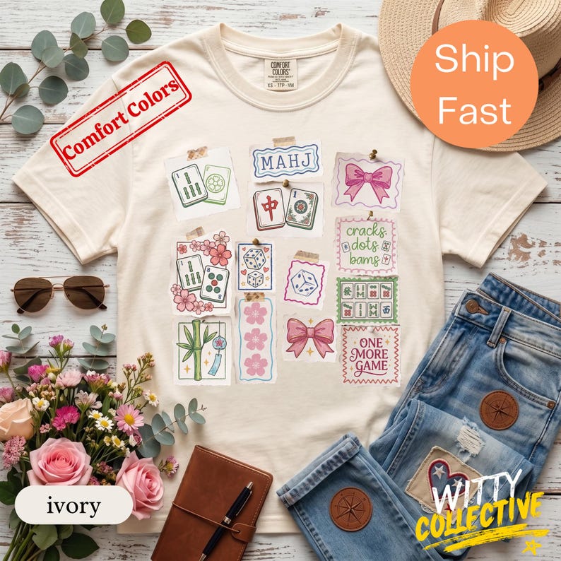 May include: Ivory-colored t-shirt with a collage of Mahjong tile illustrations, dice, and pink bows. The shirt has the text "Comfort Colors" and "ONE MORE GAME". Sunglasses, flowers, and jeans are also in the image.