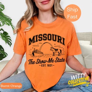 Puede incluir: Camiseta naranja quemado con un gr&aacute;fico negro que presenta la palabra "MISSOURI" sobre una ilustraci&oacute;n de un caballo, un arco y un barco. El texto "The Show-Me State - EST. 1821-" est&aacute; debajo. La camiseta tiene mangas cortas.