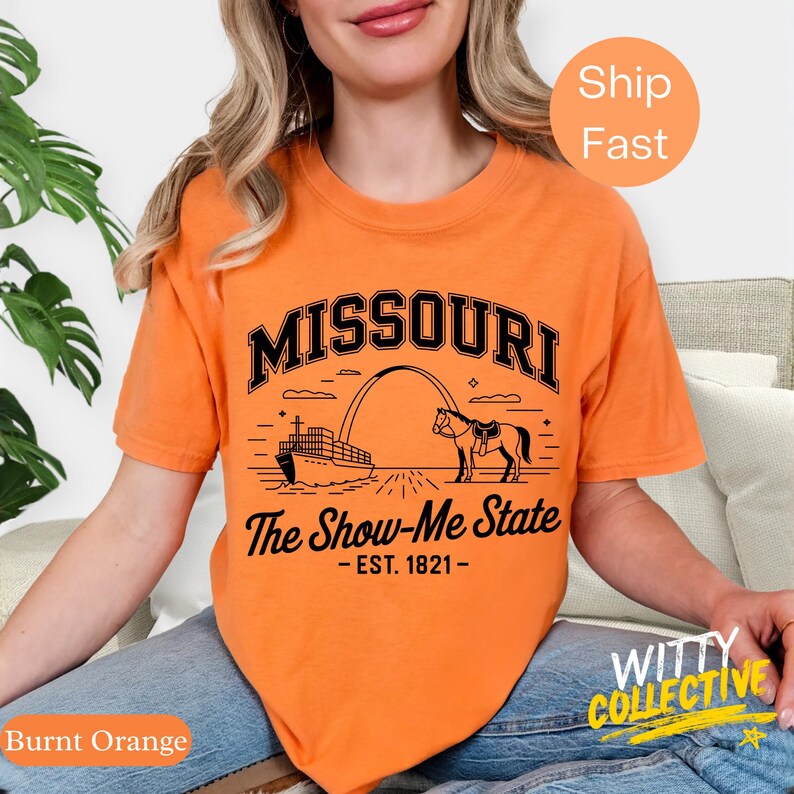 Puede incluir: Camiseta naranja quemado con un gr&aacute;fico negro que presenta la palabra "MISSOURI" sobre una ilustraci&oacute;n de un caballo, un arco y un barco. El texto "The Show-Me State - EST. 1821-" est&aacute; debajo. La camiseta tiene mangas cortas.