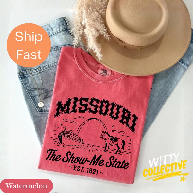 Puede incluir: Una camiseta de color sand&iacute;a con un gr&aacute;fico negro que dice "MISSOURI" encima de una imagen del Gateway Arch, un barco de vapor y un caballo. El texto "The Show-Me State - EST. 1821-" est&aacute; debajo. La camiseta est&aacute; sobre unos vaqueros azules y un sombrero beige.