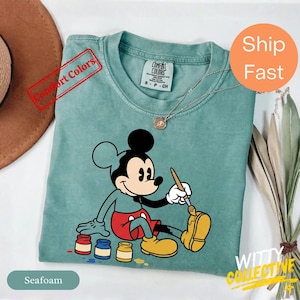 May include: Seafoam green t-shirt featuring a seated Mickey Mouse painting with a paintbrush. Mickey is wearing red shorts and yellow shoes. The shirt has a Comfort Colors tag and a gold necklace. The text "Comfort Colors" is stamped on the shirt.