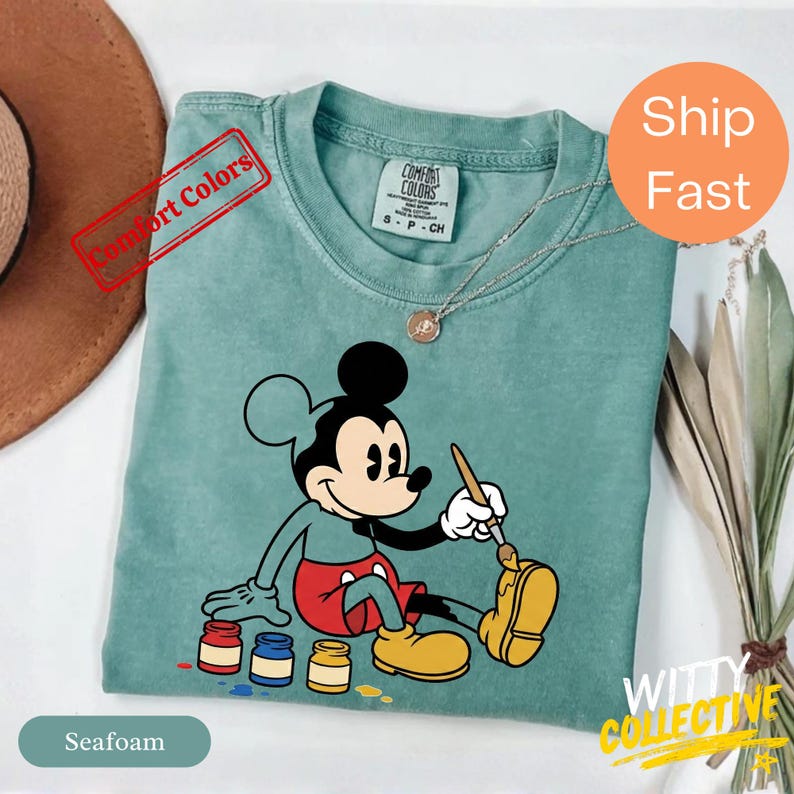 May include: Seafoam green t-shirt featuring a seated Mickey Mouse painting with a paintbrush. Mickey is wearing red shorts and yellow shoes. The shirt has a Comfort Colors tag and a gold necklace. The text "Comfort Colors" is stamped on the shirt.
