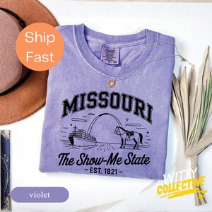 Puede incluir: Camiseta violeta con la palabra "MISSOURI" en negro, y el texto "The Show-Me State - EST. 1821-". La camiseta presenta un gr&aacute;fico de un barco, un arco y un caballo. La camiseta tiene cuello redondo y mangas cortas.