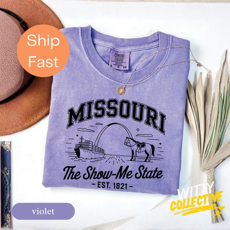 Puede incluir: Camiseta violeta con la palabra "MISSOURI" en negro, y el texto "The Show-Me State - EST. 1821-". La camiseta presenta un gr&aacute;fico de un barco, un arco y un caballo. La camiseta tiene cuello redondo y mangas cortas.