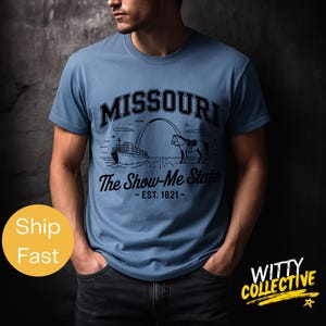 Puede incluir: Camiseta azul claro con un dise&ntilde;o gr&aacute;fico negro. El dise&ntilde;o incluye la palabra "MISSOURI" encima de una ilustraci&oacute;n de un barco, un arco y un caballo, con el texto "The Show-Me State - EST. 1821-" debajo. La camiseta la lleva una persona.