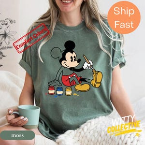 May include: A moss green t-shirt featuring a graphic of Mickey Mouse painting his yellow shoe. Mickey is wearing red shorts and yellow shoes. The shirt has the words "Comfort Colors" and "Ship Fast" printed on it.