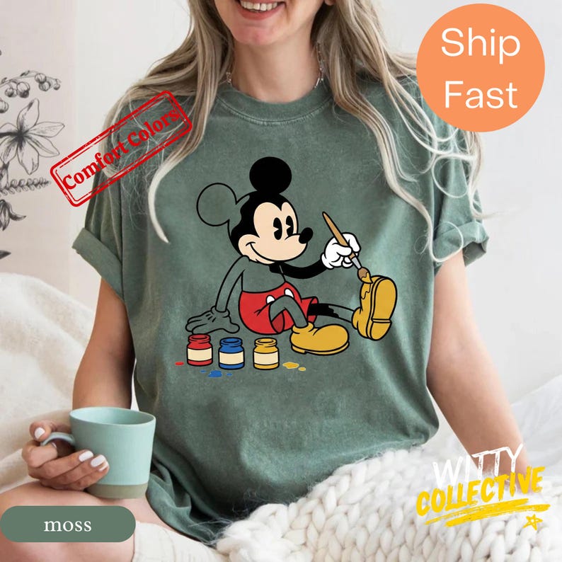 May include: A moss green t-shirt featuring a graphic of Mickey Mouse painting his yellow shoe. Mickey is wearing red shorts and yellow shoes. The shirt has the words "Comfort Colors" and "Ship Fast" printed on it.