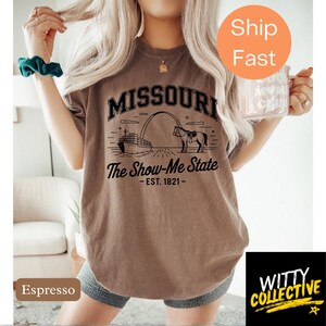 Puede incluir: Camiseta color espresso con la palabra "MISSOURI" sobre una ilustraci&oacute;n del Gateway Arch, un barco de vapor y un caballo. El texto "The Show-Me State - EST. 1821-" est&aacute; debajo. Una pegatina naranja dice "Ship Fast".
