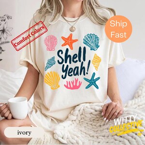 Könnte beinhalten: Elfenbeinfarbenes T-Shirt mit dem Text "Shell Yeah!" und bunten Muschelgrafiken. Das Shirt hat einen Rundhalsausschnitt und kurze Ärmel. Das Shirt ist mit "Comfort Colors" und "ivory" gekennzeichnet.