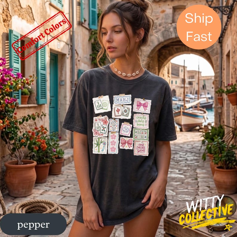 May include: Dark gray t-shirt with a collage of Mahjong tiles and pink bows. The shirt has the text "MAHJ", "crack dots & bang", and "ONE MORE GAME". The shirt is labeled "Comfort Colors" and "pepper".