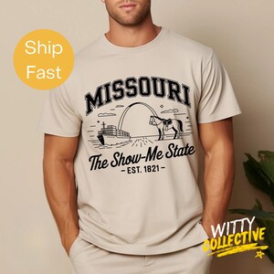 Puede incluir: Camiseta beige con un gr&aacute;fico negro del arco de Missouri, un caballo y un barco. El texto "MISSOURI" est&aacute; arqueado en la parte superior, con "The Show-Me State" y "EST. 1821" debajo. La camiseta es de color neutro.
