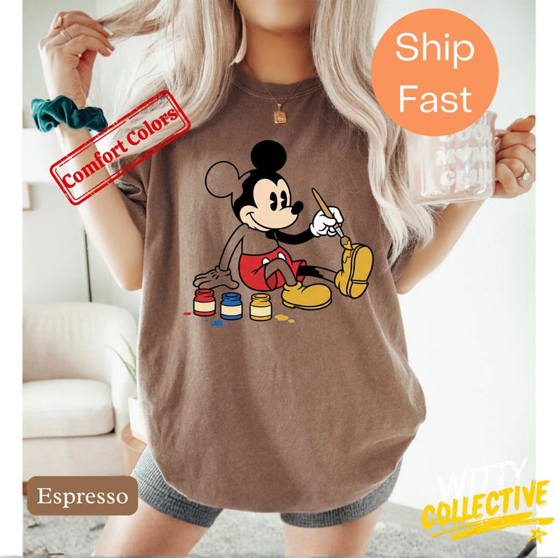 May include: Espresso-colored t-shirt featuring a graphic of Mickey Mouse painting. Mickey is depicted in his classic attire, with red shorts, yellow shoes, and white gloves. The shirt also has the text "Comfort Colors" and "Witty Collective".