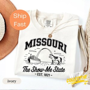 Puede incluir: Camiseta de color marfil con un gr&aacute;fico negro del arco de Missouri, un barco y un caballo. El texto "MISSOURI" est&aacute; arqueado en la parte superior, con "The Show-Me State" y "EST. 1821" debajo. Un collar dorado est&aacute; sobre la camiseta.