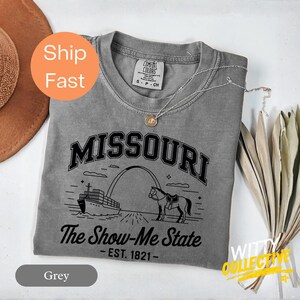 Puede incluir: Una camiseta gris Comfort Colors con un gr&aacute;fico negro de la palabra "MISSOURI" sobre una ilustraci&oacute;n de un caballo, un barco y el Gateway Arch. El texto "The Show-Me State - EST. 1821-" est&aacute; debajo. Un collar plateado est&aacute; sobre la camiseta.