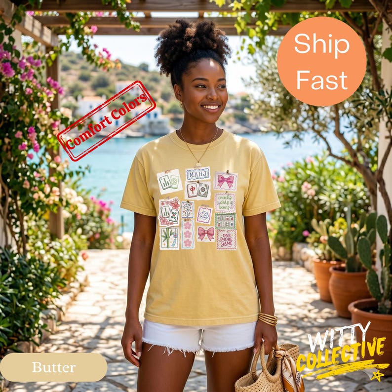 May include: A woman wearing a butter-colored t-shirt with a collage of graphic designs. The designs include images of Mahjong tiles, bows, and floral elements. The shirt is paired with white shorts. The background features a coastal scene and floral accents.