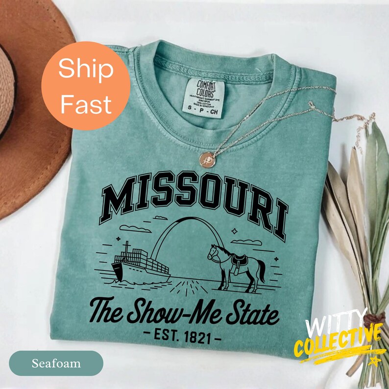 Puede incluir: Camiseta verde espuma con la palabra "MISSOURI" en letras negras. Debajo de la palabra "MISSOURI" est&aacute; el texto "The Show-Me State - EST. 1821-". La camiseta presenta un gr&aacute;fico de un barco, un arco y un caballo. Una cadena de plata est&aacute; en la camiseta.