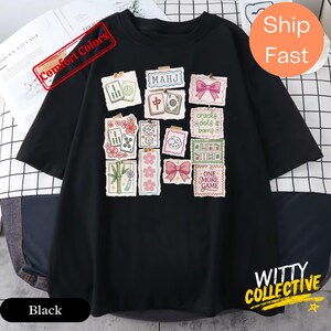 May include: Black t-shirt featuring a collage of colorful Mahjong-themed graphics, including tiles, flowers, and bows. The shirt has the text "MAHJ" and "ONE MORE GAME". The shirt is from the "WITTY COLLECTIVE" and has "Comfort Colors" printed on it.