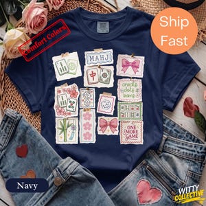 May include: Navy blue t-shirt featuring a collage of Mahjong tile illustrations. The design includes text that reads "MAHJ", "cracks dots & bams", and "ONE MORE GAME". The shirt is paired with denim jeans.