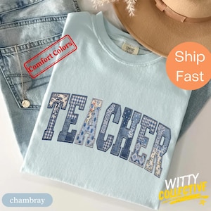 May include: A pale blue t-shirt featuring the word "TEACHER" in a patchwork design of blue and white patterns. The shirt is folded on top of a pair of blue denim jeans.