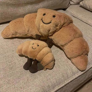 May include: Two plush croissant toys in shades of brown. The larger croissant has a smiling face and brown legs. The smaller croissant also has a smiling face and brown legs. The toys are on a gray surface.
