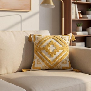 May include: A decorative throw pillow with a geometric design in yellow and white. The pillow features a textured pattern and is adorned with tassels at each corner. The pillow is placed on a beige sofa, suggesting a home decor theme.