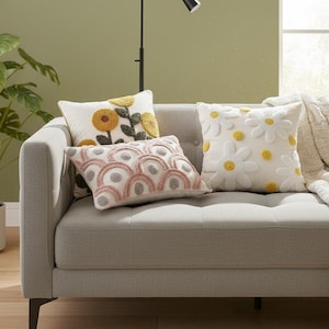 Peut inclure: Un canapé gris avec trois coussins décoratifs. Un coussin présente un motif de tournesol jaune, un autre un motif en arc rose et gris, et le troisième des marguerites blanches avec des centres jaunes.