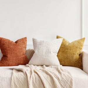 May include: Three decorative throw pillows in shades of orange, white, and gold rest on a white sofa. The pillows have textured surfaces. A cream-colored throw blanket with a diamond pattern is draped over the sofa.