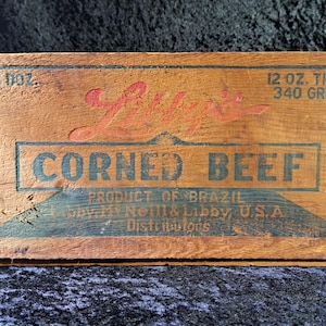 May include: A vintage wooden crate with the words "Luy's CORNED BEEF" in red and blue lettering. The crate also has text indicating the product is from Brazil and distributed by Libby, U.S.A. The crate is marked "12 OZ. TINS 340 GRM."