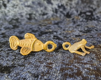 May include: Three small, detailed metal charms in the shape of a fish, frog, and lizard. The charms are gold-toned and have a loop for attaching to jewelry or other crafts. The background is a dark, textured fabric.