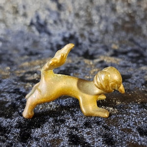 May include: A small, bronze-colored cat figurine. The cat is in a walking pose with its tail raised. The figurine has a smooth, polished finish and is set against a dark, textured background.