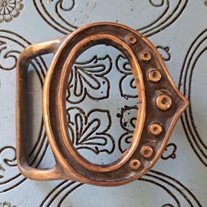 May include: A vintage copper-colored belt buckle with an oval shape and a rectangular bar. The buckle has decorative circular accents along one side. The background is a light blue surface with a floral pattern.