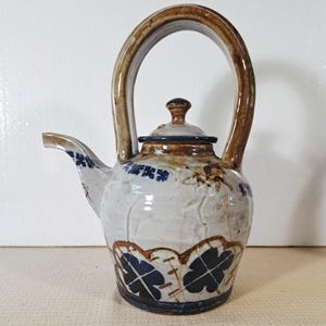 May include: A ceramic teapot with a high arched handle and a short spout. The body features a light gray glaze with brown and blue hand-painted floral patterns near the base and rim. It includes a matching lid with a small knob handle.