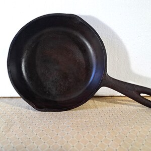 May include: A dark brown cast iron skillet with a long handle and a small pouring spout. The skillet is round and appears well-seasoned, ready for cooking. The handle has a hole for hanging.