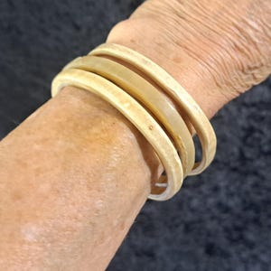 May include: Four cream-colored bangles stacked on a wrist. The bangles have a smooth, rounded design with a slightly off-white hue. The bracelets are made of a light-colored material.