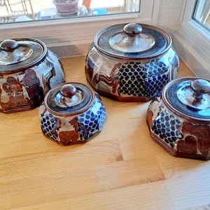 May include: A set of four octagonal ceramic canisters with lids, featuring a multi-colored glaze in shades of brown, blue, and cream. The surface displays a repeating scale-like pattern alongside abstract brown and cream patches on each container.