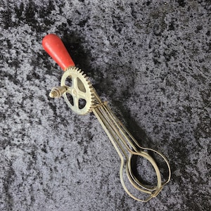 May include: A manual rotary egg beater featuring a metallic silver finish, a gear-driven mechanism, and a smooth red handle. The kitchen tool consists of a central crank wheel connected to two wire beaters designed for mixing ingredients.