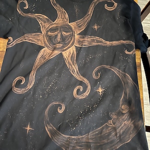 Puede incluir: Camiseta negra con un diseño celestial. El diseño incluye un sol con una cara y una luna creciente, ambos en color marrón. El fondo está lleno de pequeños detalles en forma de estrella.