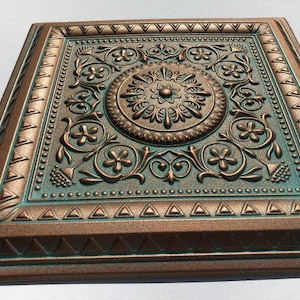 May include: Square, bronze-coloured decorative ceiling tile with an ornate floral and scroll design. The tile has a central medallion and a teal-coloured background, with a raised relief pattern and a textured finish. The edges are framed with a geometric border.