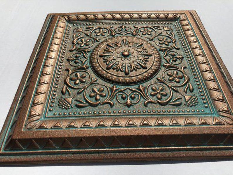 May include: Square, bronze-coloured decorative ceiling tile with an ornate floral and scroll design. The tile has a central medallion and a teal-coloured background, with a raised relief pattern and a textured finish. The edges are framed with a geometric border.