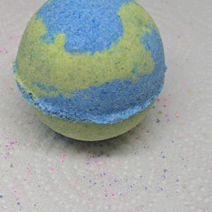 Puede incluir: Una bomba de baño esférica con una combinación de colores azul y amarillo. La bomba de baño tiene una superficie texturizada y granulada. Los colores están mezclados, creando un efecto veteado. La bomba de baño está sobre una superficie blanca.