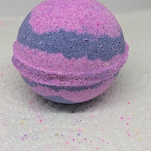 May include: A round bath bomb with a textured surface. The bath bomb features a layered design of pink and purple hues. The center band is a darker shade of purple. Small colorful particles are scattered around the bath bomb.