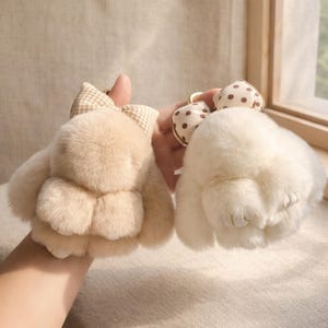May include: Two plush bunny keychains. One is beige with a plaid bow, the other is white with a polka dot bow. Both have a fluffy texture and gold-colored key rings. The bunny keychains are a cute accessory.