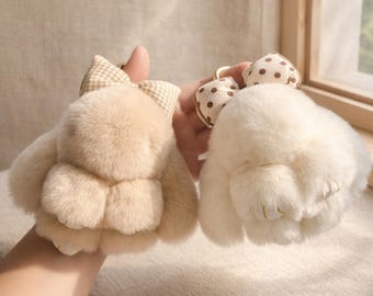 May include: Two plush bunny keychains. One is beige with a plaid bow, the other is white with a polka dot bow. Both have a fluffy texture and gold-colored key rings. The bunny keychains are a cute accessory.