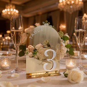 May include: A gold-trimmed table number 3 with a mirrored surface, arched top, and the word "TABLE" at the top. The number is white with a silver outline. The table setting includes champagne flutes, candles, and floral arrangements.
