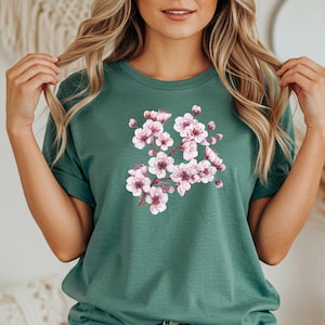 Könnte beinhalten: Ein grünes T-Shirt mit einem floralen Design aus rosa Kirschblüten. Das kurzärmelige Oberteil ist aus weichem Material gefertigt und wird mit blauen Jeans kombiniert. Die florale Grafik verleiht dem lässigen Outfit einen Hauch von Natur.