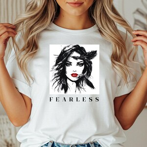 Könnte beinhalten: Weißes T-Shirt mit einer schwarz-weißen Grafik eines Frauengesichts mit rotem Lippenstift und einer Feder. Das Wort "FEARLESS" ist darunter in Schwarz gedruckt. Das T-Shirt ist aus weichem Stoff.