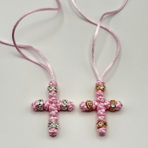 May include: Two pink cross necklaces with decorative silver and gold accents. The crosses are made of woven pink material and feature sparkling embellishments. Each necklace has a matching pink cord.