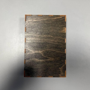 May include: A rectangular wooden box with a dark gray finish and visible wood grain. The corners and edges feature a contrasting light brown color, with small rectangular cutouts. The box is empty and sits on a white surface.
