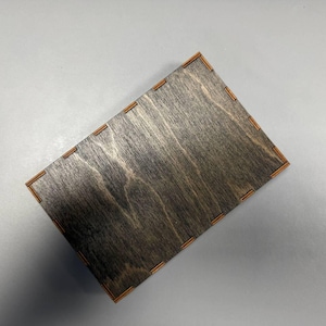 May include: A rectangular wooden box with a dark, stained finish. The box has a natural wood grain pattern and is accented with lighter-colored wood at the corners. The box is empty and sits on a gray surface.