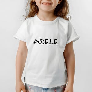 K&ouml;nnte beinhalten: Wei&szlig;es T-Shirt mit dem Namen "ADELE" in schwarzer, handschriftlicher Schrift. Das Shirt hat kurze &Auml;rmel und einen Rundhalsausschnitt. Das Model tr&auml;gt eine Hose mit Muster.